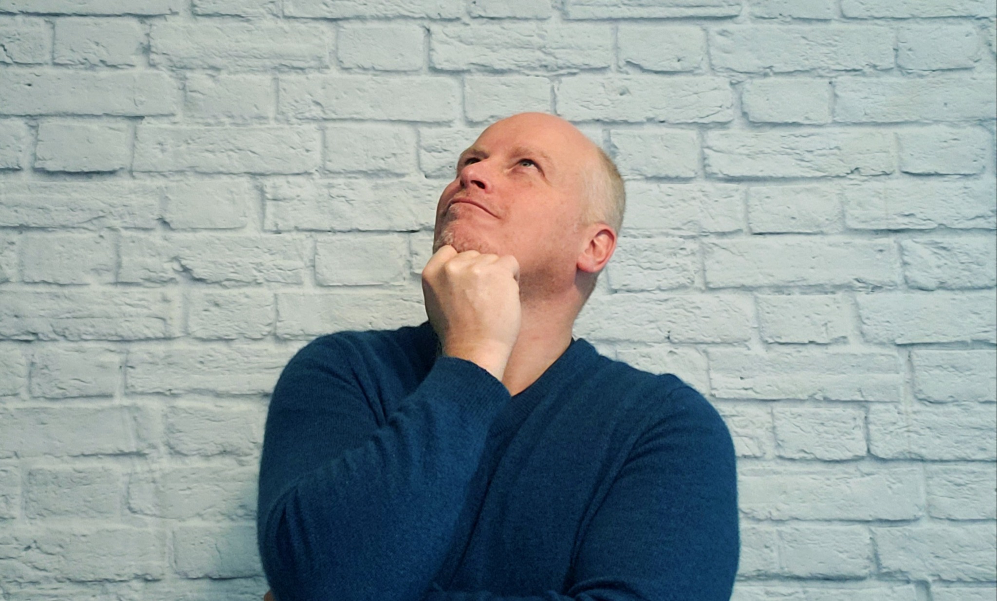 Doug Greathouse headshot - man standing infront of brick wall looking up with hand under his chin