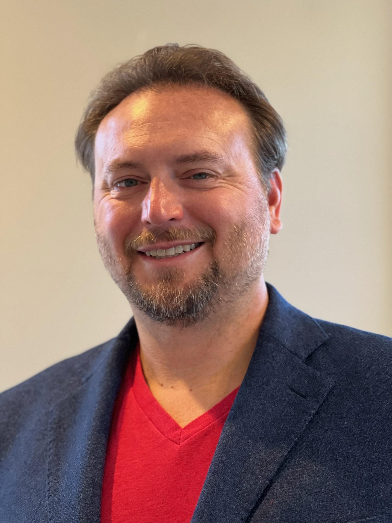 Headshot of Jason Reid - male wearing navy suit with red shirt smiling